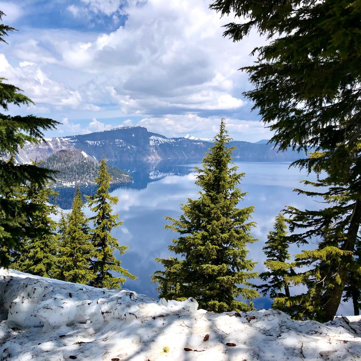 A mostly cloudy day, reflections of clouds in the lake, snow lingering around the caldera rim. 
