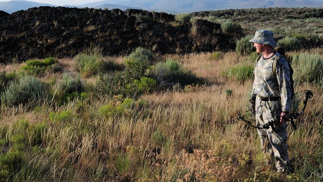 a man in camouflage with a bow standing in an open grassy field