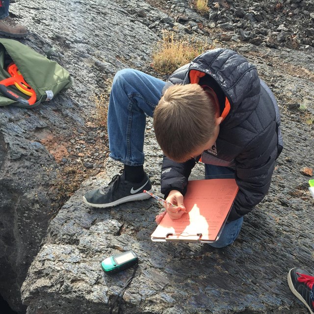 a boy writing on a clipboard crouching on the ground