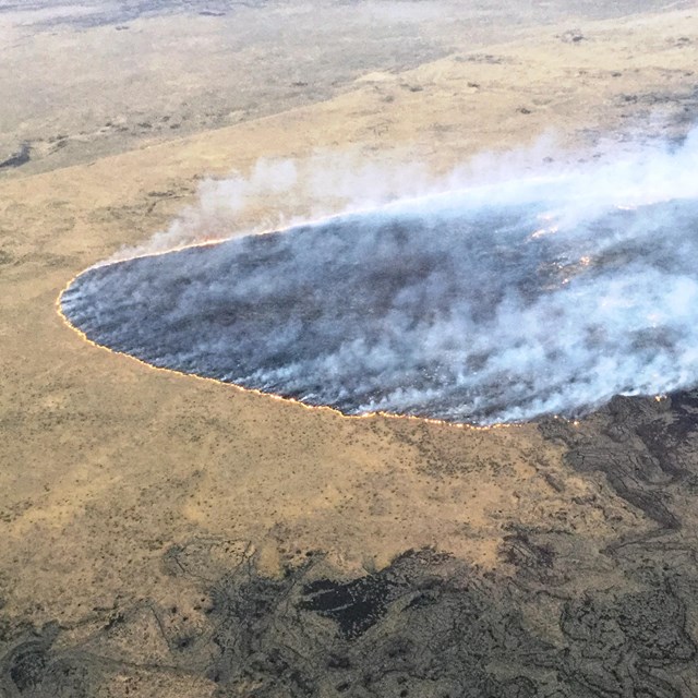 aerial view of a fire burning a grassland on the edge of an area of dark rocks