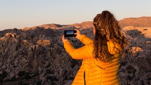 Girl in orange jacket takes picture with smart phone of rocky landscape