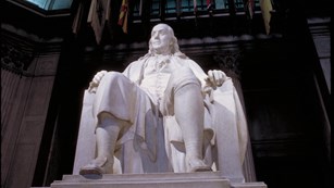 White, seated statue of Benjamin Franklin in darkened globed room.