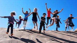 Group of children jump over large sand dune with blue sky in background.