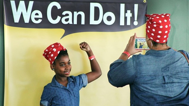 Visitor taking a picture of a kid wearing a Rosie the Riveter bandana by a You Can Do It frame