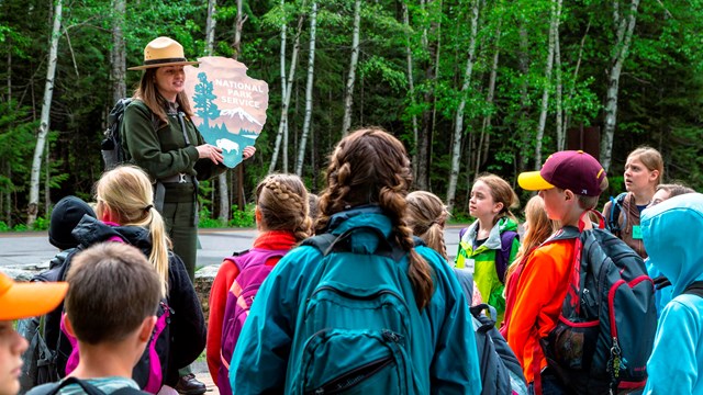 Ranger holding an NPS arrowhead logo while talking to a school group outside