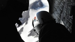 Crew member wearing a helmet peers out of a helicopter down on a mountain