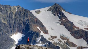 Hiker on a rocky outcropping near the top of a snow-capped mountain