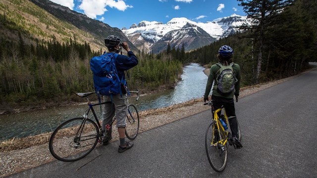 Two bicyclists wearing helmets and backpacks stopped on a road to view a mountain
