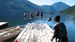 Person standing on a dock of a lake watching four other people jump in
