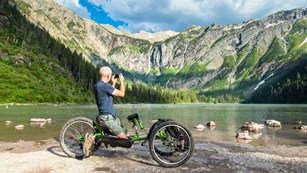 Man in wheelchair along edge of lake taking picture of mountain scene