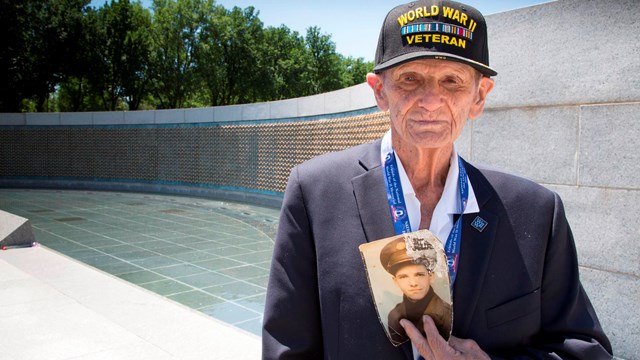 Older veteran holding a photo of himself as a soldier while standing in front of a memorial pond