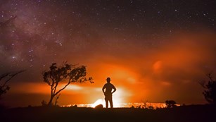 Silhouette of a ranger in front of a lava eruption, trees, and night sky