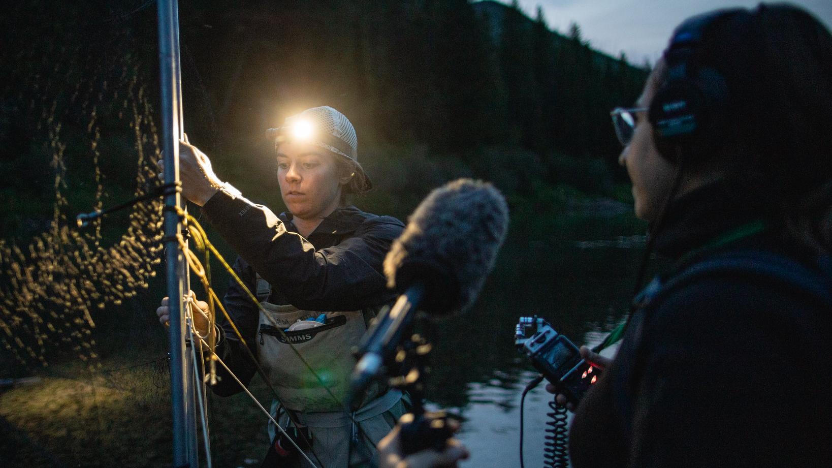 A person with headphones and a microphone stands near a person wearing a headlamp in front of a net