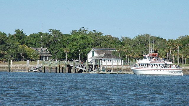 A white ferry boat carrying passengers approaching the dock of a tree covered island.