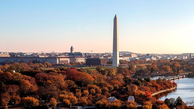 The Washington Monument obelisk rises above autumn trees, a blue river, and the low city beyond.