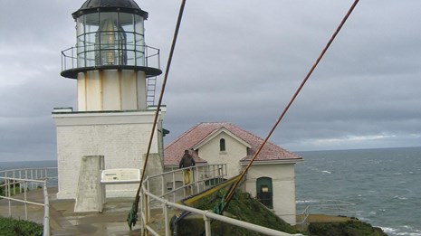 A lighthouse with a squat tower on a bluff overlooking an expanse of water under overcast sky