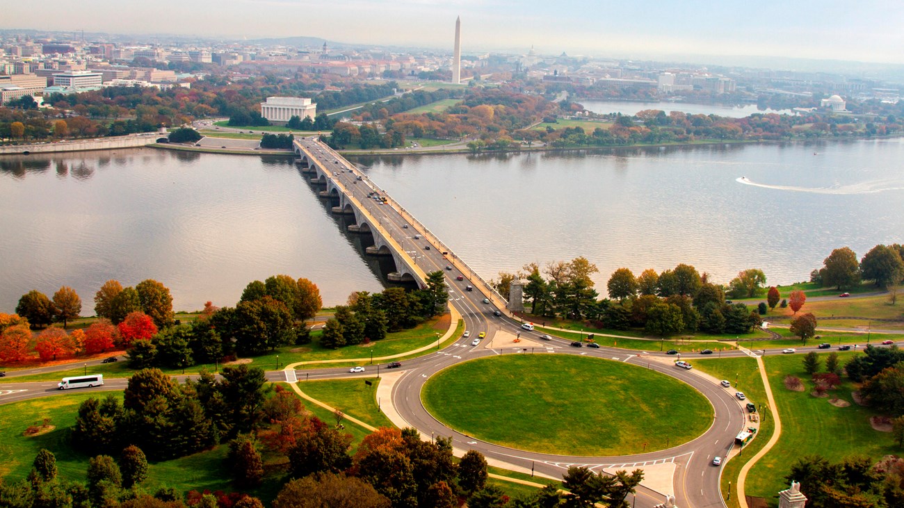 Cars move around a traffic circle and bridge over a river, toward a city with trees and monuments