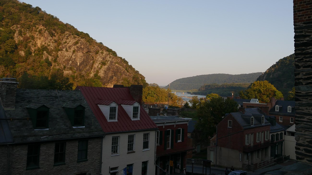 Two-story brick structures line a road, with a river and tree-covered hillsides in the background