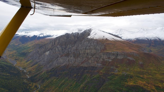 A thin ribbon of water winds through a valley below snow tipped mountains, seen from a plane