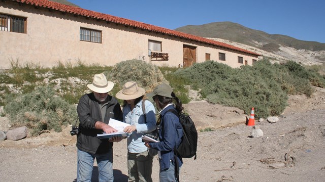 Three people equipped for field work gather over a report in a dry southern California landscape