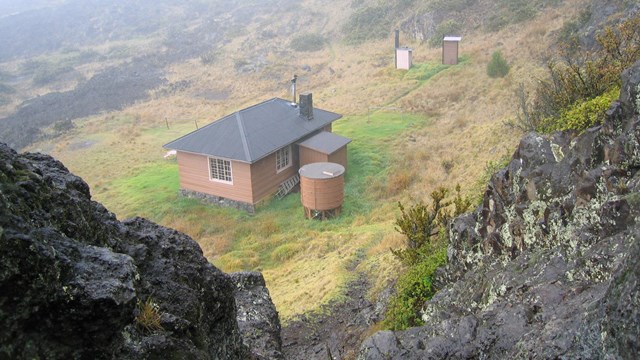 View through dark, lichen-covered rocks to a rectangular structure and water cistern in a clearing