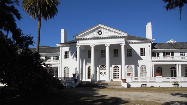 A driveway curves in front of a large white estate home with four tall columns and many windows,