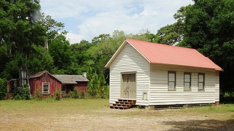 A single-story structure in a clearing has a door at one end and windows along the side.
