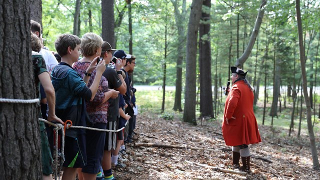 An interpreter dressed as a British soldier in a red coat talks to crowd of people in a wooded area