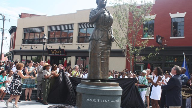 A crowd of people in a city plaza pull back a sheet to unveil a tall statue of Maggie Lena Walker