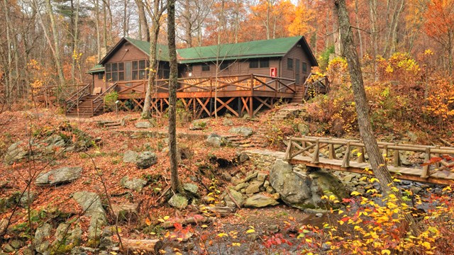 A rustic camp in a wooded area with a wooden bridge, stone steps. Deciduous trees have fall foliage.