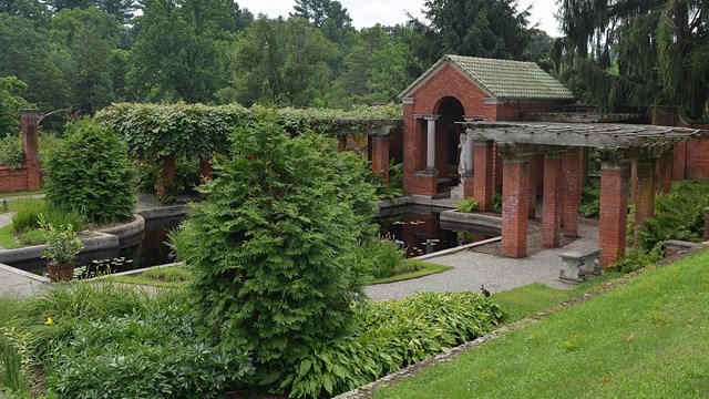 A brick pool house and pergola in a formal garden, beside a pond, garden beds, and terracing