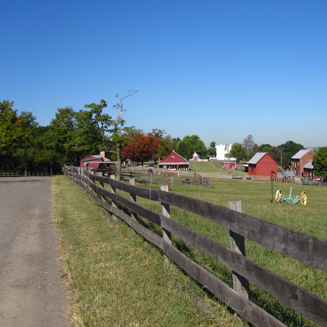 A wooden fence runs along a road that leads towards a cluster of farm buildings.