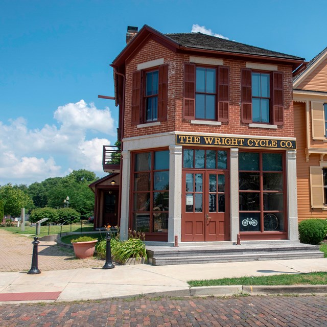 A two-story shop on a brick road has large pane glass windows with a sign above: The Wright Cycle Co
