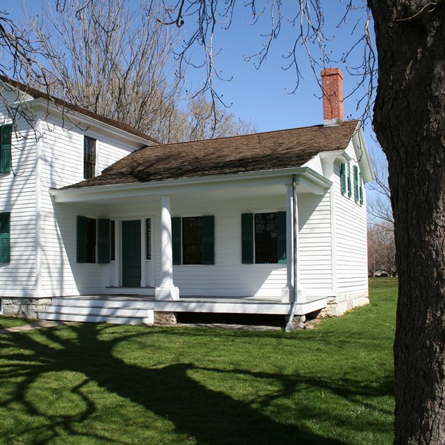 A two story white house with a single story ell on a neat lawn under the shadow of a tall tree