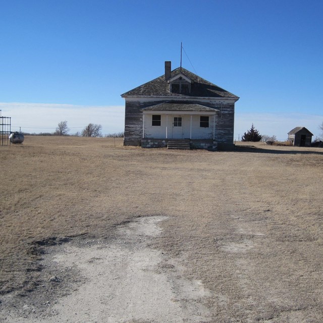 A small wooden schoolhouse on a flat expanse of short, dry grass under blue sky.