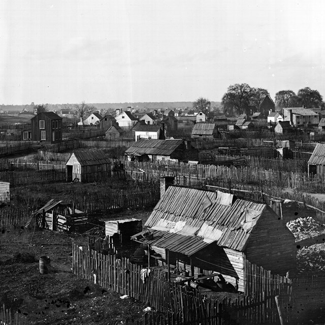 Wooden fencing winds around a collection of small wooden houses on a flat landscape, b & w photo