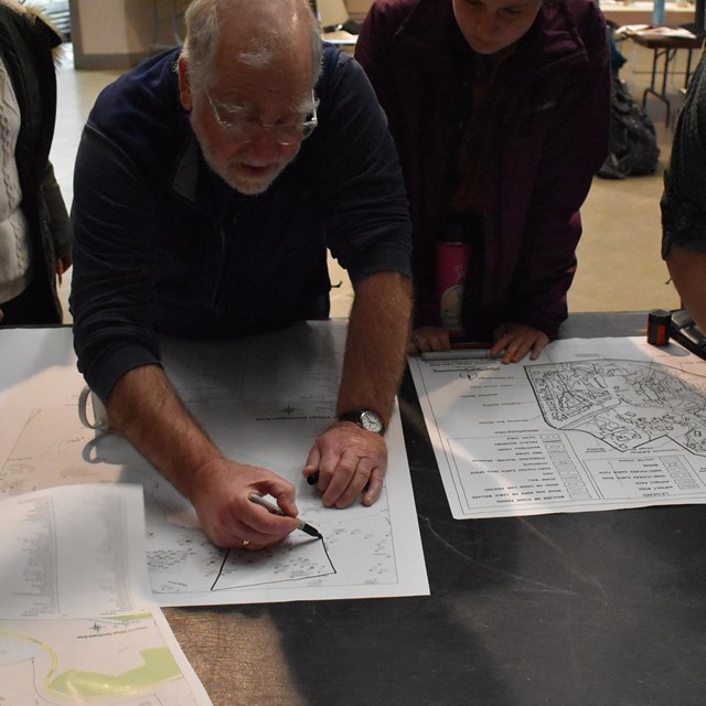 Students gather and watch as a man leans over a table and draws on papers spread on its surface 