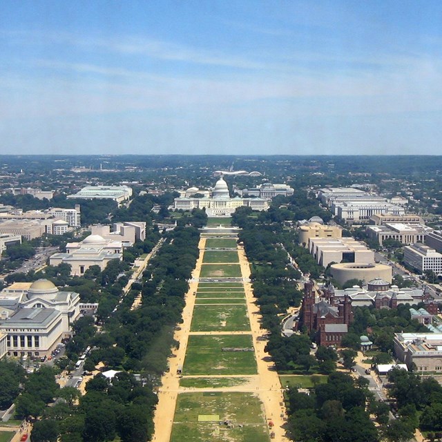 Aerial view of worn turf on the National Mall, surrounded by buildings and trees 