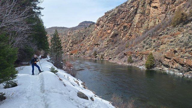 Person wearing winter gear and snowshoes stands on a snow-covered trail along a flowing reservoir.