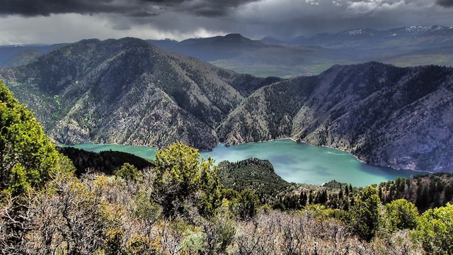 A deep canyon with a blue-green reservoir surrounded by steep brushy slopes and a cloudy sky.