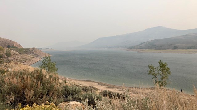 Hazy wildfire smoke over a large blue reservoir. Sagebrush vegetation in foreground. Mesas far off.