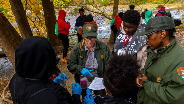 Students and two uniformed rangers stand outside wearing blue gloves and looking down at a graph.