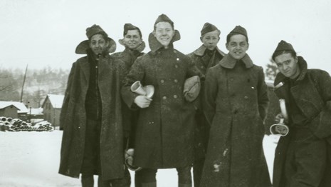 Black and white photo of six smiling young men standing out in the snow, wearing long wool coats.
