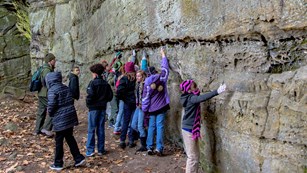 Uniformed ranger and group of students investigate a green-and-turquoise streaked, craggy rock wall.