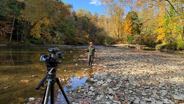 Uniformed ranger stands in front of a video camera in water near the edge of a river.