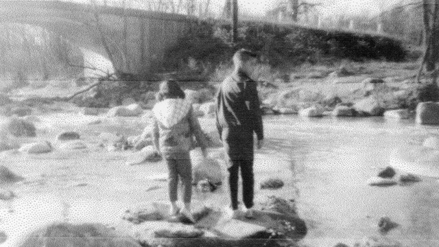 Blurry black and white photo of two children standing on boulders in a river.