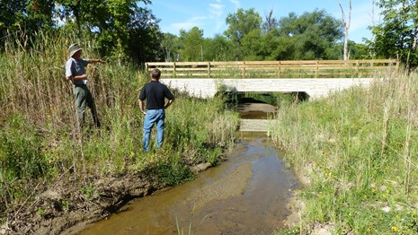 Uniformed ranger stands next to small creek with another person, pointing toward concrete culvert.