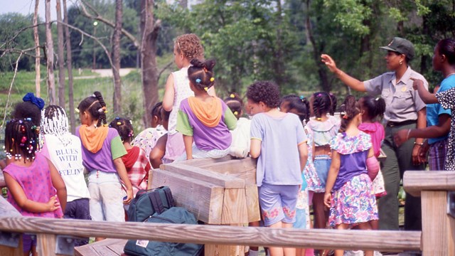 A black, female ranger provides a tour to a school group of children at a marsh.
