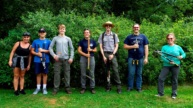 Seven smiling people stand in a line in front of green shrubs holding branch cutting tools.