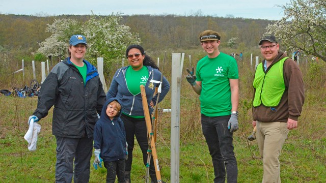 Five smiling people stand in a field next to a white vertical tube and a small tree in a bucket.